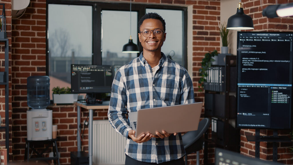 Portrait of african american cloudsecurity programer standing working on laptop looking up and smiling